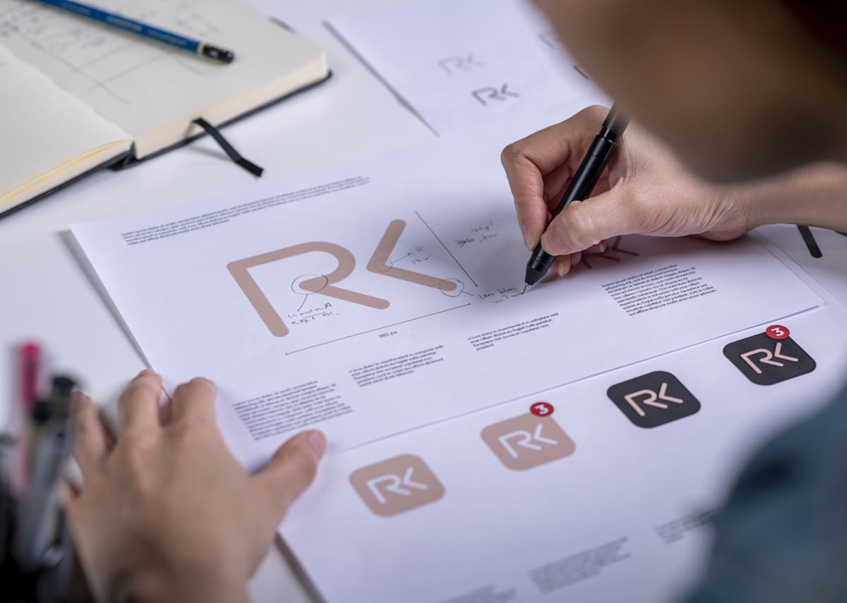 A person works on designing a logo, drawing precise details on a printout featuring a stylized 'RK' logo. The desk is scattered with design drafts, including color variations and notes. A notebook and a pen are visible in the background, emphasizing the creative process involved in developing a strong brand identity.