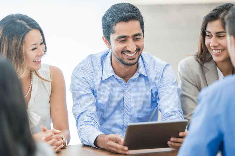 Group of professionals smiling and discussing work around a tablet during a meeting, representing collaboration and communication in science marketing.